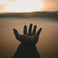 Das Bild zeigt eine ausgestreckte Hand vor einem See und Sonnenuntergang.